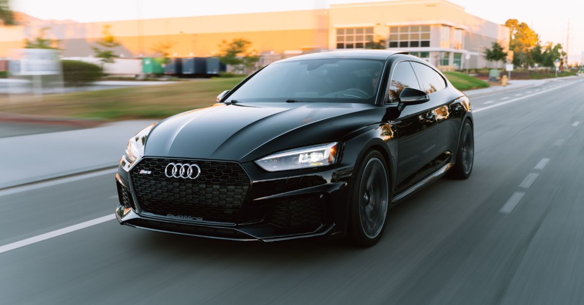 Black Audi RS5 Sportback driving on a smooth city road at sunset, featuring LED headlights, black alloy wheels, and a sleek sporty design with motion blur in the background.