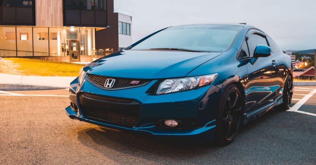Blue Honda Civic Si coupe parked in a modern parking lot, featuring sporty styling, black wheels, and an aggressive front grille.