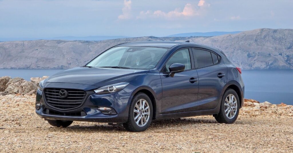 Blue Mazda3 hatchback parked on a coastal overlook, featuring sleek styling, alloy wheels, and a scenic ocean backdrop.