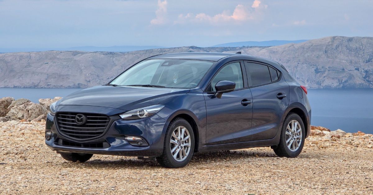 Blue Mazda3 hatchback parked on a coastal overlook, featuring sleek styling, alloy wheels, and a scenic ocean backdrop.