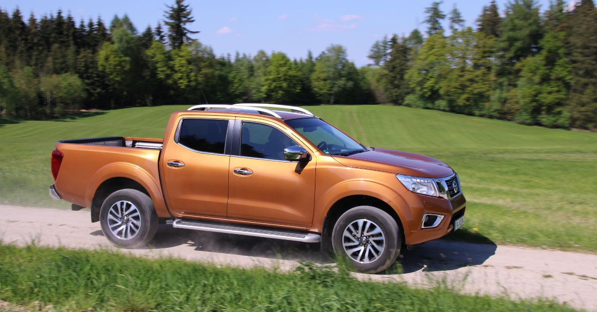 Orange Nissan Navara pickup truck driving on a dirt road, featuring rugged styling, alloy wheels, and off-road capability.