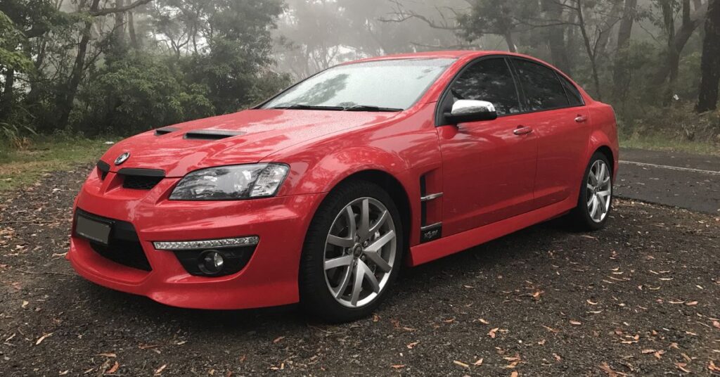 Red Holden Commodore SS V sedan parked in a misty forest, featuring sporty styling, alloy wheels, and an aggressive front design.