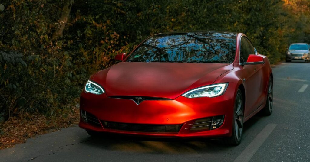Red Tesla driving on a tree-lined road, featuring sleek styling, LED headlights, and a premium all-electric design.