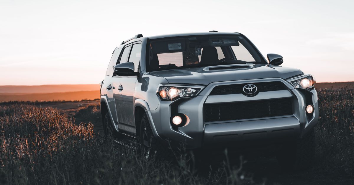Silver Toyota 4Runner SUV with headlights illuminated, parked in a grassy field during sunset, with a wide open landscape and warm evening sky in the background.