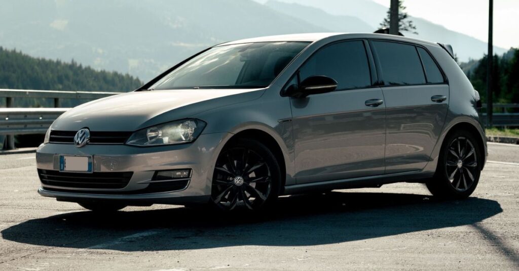 White Volkswagen Golf hatchback with black alloy wheels parked on a paved roadside, set against a scenic mountain landscape under a bright sky.